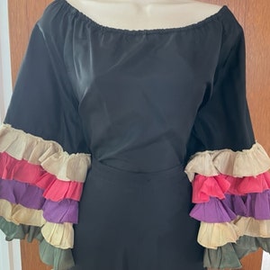 Puede incluir: Vestido negro de hombros descubiertos con mangas de volantes. Las mangas están superpuestas con volantes en crema, rosa, morado y verde.