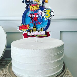 K&ouml;nnte beinhalten: Ein wei&szlig;er Kuchen mit einem Spiderman-Motiv-Topper. Der Topper zeigt Spiderman, Spider-Gwen und andere Figuren aus der Zeichentrickserie Amazing Friends. Der Topper enth&auml;lt auch die Worte "POW!" und "BAM!" sowie den Namen "LANDON AMAZING FRIENDS".