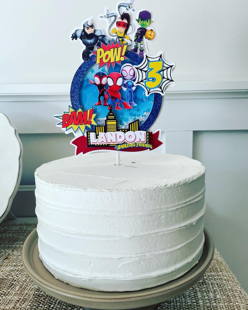 K&ouml;nnte beinhalten: Ein wei&szlig;er Kuchen mit einem Spiderman-Motiv-Topper. Der Topper zeigt Spiderman, Spider-Gwen und andere Figuren aus der Zeichentrickserie Amazing Friends. Der Topper enth&auml;lt auch die Worte "POW!" und "BAM!" sowie den Namen "LANDON AMAZING FRIENDS".
