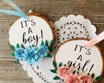 May include: Two round wood slice ornaments with white painted centers. One features the text "IT'S A boy!" with light blue paper flowers and green glitter leaves. The other features "IT'S A girl!" with light pink paper flowers and green glitter leaves.