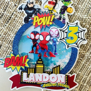 K&ouml;nnte beinhalten: Ein blauer und wei&szlig;er Tortendekor mit Spiderman-Motiv. Es zeigt einen roten und blauen Spiderman, einen schwarzen und roten Spiderman, einen wei&szlig;en und rosafarbenen Spiderman, eine gr&uuml;ne und violette Figur, eine gelbe und schwarze Figur, eine gelbe Zahl 3 und die Worte "BAM!" und "POW!" Der Dekor enth&auml;lt auch ein rotes Banner mit den Worten "LANDON'S AMAZING FRIENDS".