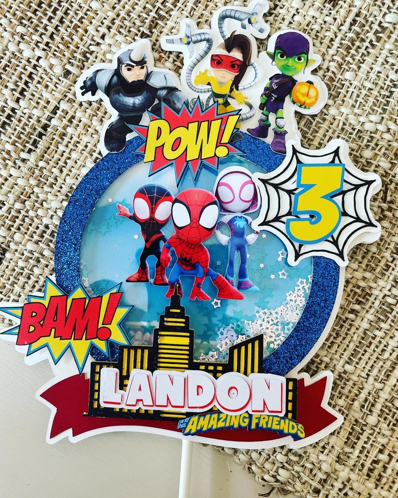 K&ouml;nnte beinhalten: Ein blauer und wei&szlig;er Tortendekor mit Spiderman-Motiv. Es zeigt einen roten und blauen Spiderman, einen schwarzen und roten Spiderman, einen wei&szlig;en und rosafarbenen Spiderman, eine gr&uuml;ne und violette Figur, eine gelbe und schwarze Figur, eine gelbe Zahl 3 und die Worte "BAM!" und "POW!" Der Dekor enth&auml;lt auch ein rotes Banner mit den Worten "LANDON'S AMAZING FRIENDS".
