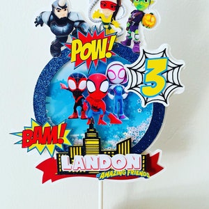 K&ouml;nnte beinhalten: Ein Tortenaufsatz mit Spider-Man und seinen fantastischen Freunden. Der Aufsatz ist mit einem blauen und wei&szlig;en Hintergrund, Sternen und den Worten "POW!" und "BAM!" verziert. Der Aufsatz enth&auml;lt auch eine Skyline, ein rotes Band und den Text "LANDON'S AMAZING FRIENDS".