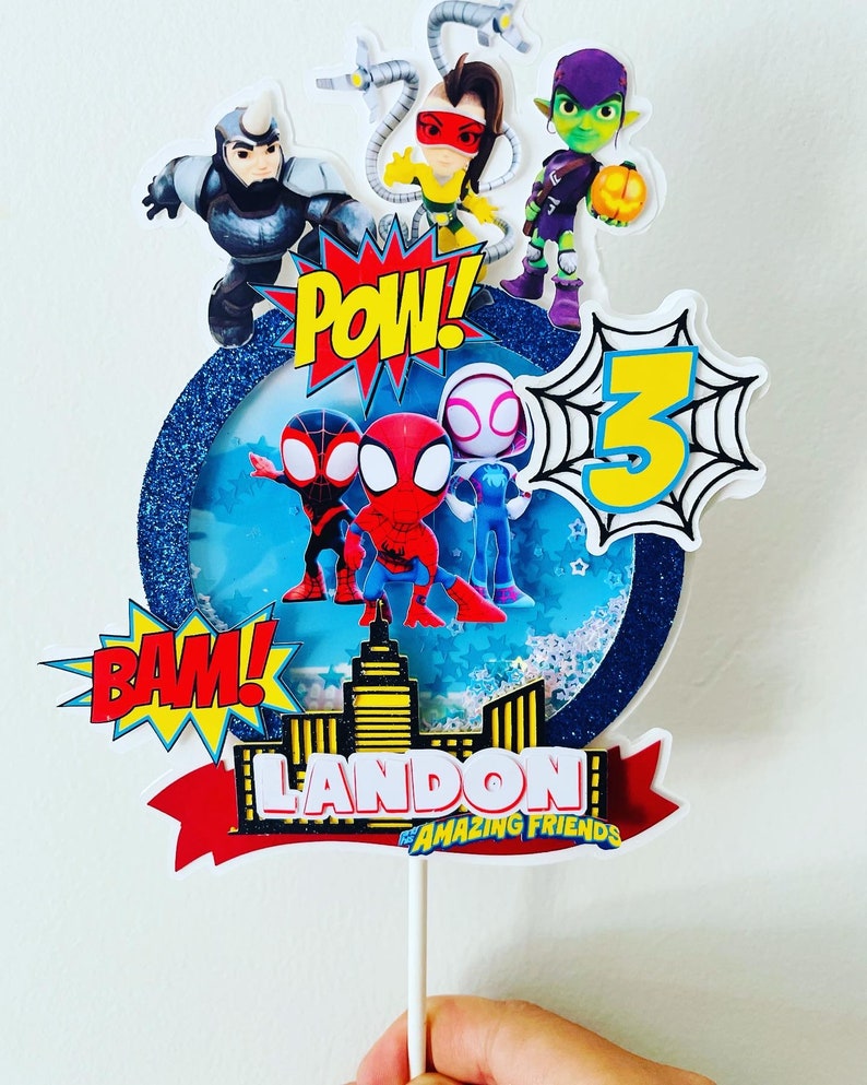 K&ouml;nnte beinhalten: Ein Tortenaufsatz mit Spider-Man und seinen fantastischen Freunden. Der Aufsatz ist mit einem blauen und wei&szlig;en Hintergrund, Sternen und den Worten "POW!" und "BAM!" verziert. Der Aufsatz enth&auml;lt auch eine Skyline, ein rotes Band und den Text "LANDON'S AMAZING FRIENDS".