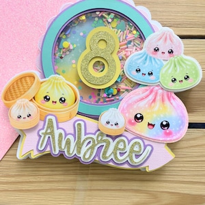 May include: A circular shaker cake topper featuring a teal border, a gold glitter number 8, and colorful confetti inside. It is decorated with multiple kawaii-style dumpling illustrations and a banner with the name Aubree in gold glitter script.