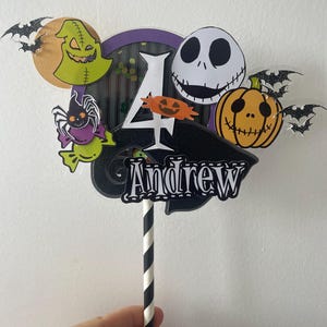Op de afbeelding: Een kleurrijke taarttopper met personages uit The Nightmare Before Christmas. Het ontwerp bevat Jack Skellington, een pompoen en de naam "Andrew" met het nummer "4". De topper wordt vastgehouden door een zwart-wit gestreepte rietje.