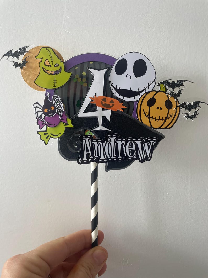Op de afbeelding: Een kleurrijke taarttopper met personages uit The Nightmare Before Christmas. Het ontwerp bevat Jack Skellington, een pompoen en de naam "Andrew" met het nummer "4". De topper wordt vastgehouden door een zwart-wit gestreepte rietje.