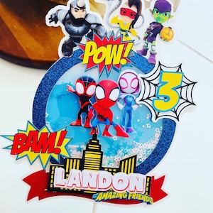 K&ouml;nnte beinhalten: Ein blauer und wei&szlig;er Tortendekor mit Spiderman-Motiv. Es zeigt Spiderman und seine Freunde, eine Stadtlandschaft und die Worte "Landon's Amazing Friends" sowie "Pow!" und "Bam!" im Comic-Stil.
