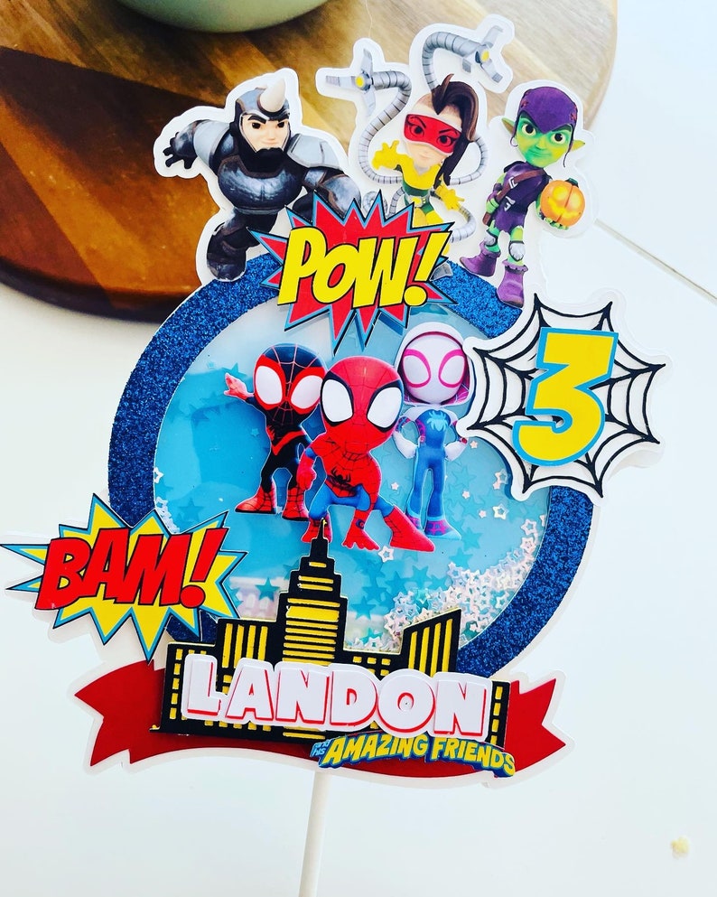 K&ouml;nnte beinhalten: Ein blauer und wei&szlig;er Tortendekor mit Spiderman-Motiv. Es zeigt Spiderman und seine Freunde, eine Stadtlandschaft und die Worte "Landon's Amazing Friends" sowie "Pow!" und "Bam!" im Comic-Stil.