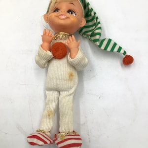 May include: A doll with blonde hair wearing a white ribbed long-sleeve outfit, a green and white striped pointed hat with an orange pom-pom, and red and white striped shoes. The doll has an orange pom-pom on its chest and gold trim at the neck.