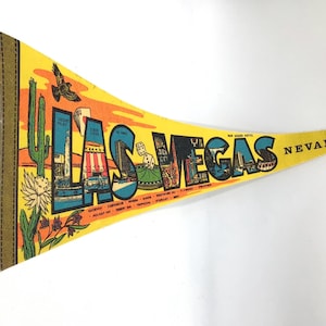 Op de afbeelding: Gele vilten wimpel met de woorden "LAS VEGAS NEVADA" in zwarte en blauwe letters. De wimpel toont illustraties van cactussen, gebouwen en een vogel in vlucht. De wimpel heeft gele lipjes om op te hangen.