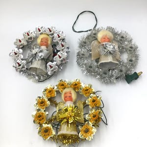 Può includere: Tre addobbi per la cima dell'albero a forma di angelo vintage con funzioni di illuminazione. Ogni addobbo presenta un angelo di plastica con capelli biondi e un vestito bianco, circondato da una ghirlanda di stelle filanti e fiori. Le ghirlande sono argentate, dorate e gialle.