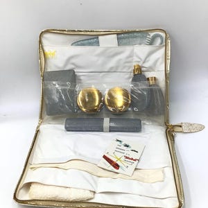 May include: A gold-colored travel case with a zipper closure. The case is open and contains a variety of toiletries, including a comb, a bottle of perfume, a small mirror, and a bar of soap. The case also includes a white towel and a small card that reads "The modern traveler is a "Diel" traveler."