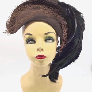 May include: A brown felt hat with a black feather and netting. The hat is angled to the side and the feather is long and flowing.