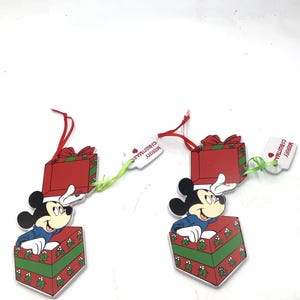 Puede incluir: Dos adornos navideños planos que muestran a Mickey Mouse saliendo de una caja de regalo verde y roja con patrones de acebo. Cada adorno tiene una tapa roja, una cinta roja para colgar y una pequeña etiqueta blanca que dice Merry Christmas.