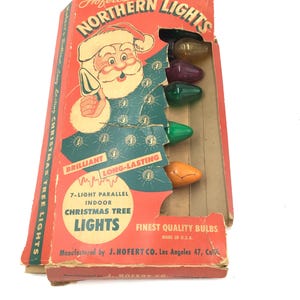 Może przedstawiać: Winietkowe pudełko z lampkami choinkowymi Hofert's Northern Lights, 7-światłowe, równoległe, do użytku wewnętrznego. Pudełko przedstawia rysunkowego Świętego Mikołaja i ekspozycję kolorowych żarówek. Na pudełku widnieje napis "Brilliant Long-Lasting" i "Finest Quality Bulbs Made in U.S.A."