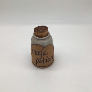 May include: A small, ceramic jar with a cork stopper. The jar is light grey with a brown label that says "magic potion".