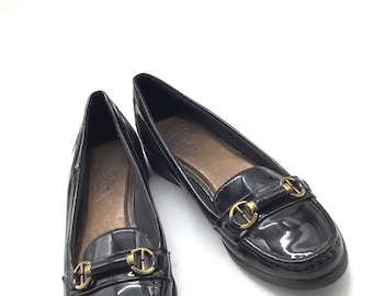 May include: A pair of black patent leather loafers featuring a rounded toe, a low heel, and a decorative gold-tone metal horsebit detail across the vamp. The interior of the shoes is lined with a dark brown material and includes a visible brand logo.