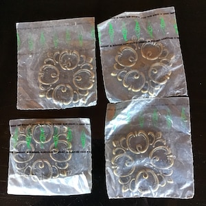 Op de afbeelding: Vier goudkleurige metalen decoratieve accenten, elk afzonderlijk verpakt in een plastic zakje. Elk accent heeft een gedetailleerd, versierd ontwerp met een bloemmotief.