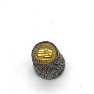 May include: A silver thimble with a gold-colored top featuring the text "U.S.S. Arizona Memorial Pearl Harbor Hawaii".