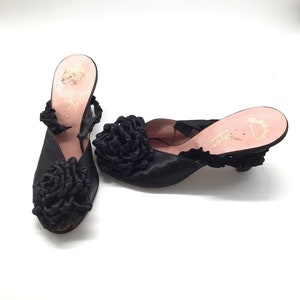 Op de afbeelding: Een paar zwarte satijnen schoenen met hak en een grote zwarte satijnen bloem op de neus. De schoenen hebben een roze voering en de tekst "Chateau Rose" is zichtbaar aan de binnenkant van de schoen.