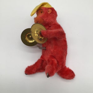 May include: A red plush monkey toy with a yellow hat plays a pair of cymbals. The monkey is sitting on a white surface.