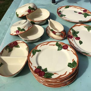 Puede incluir: Una colección de vajilla de cerámica de color crema con un diseño de frutas y ramas. El juego incluye platos, cuencos y fuentes. Los bordes son marrones y la fruta es roja con hojas verdes.