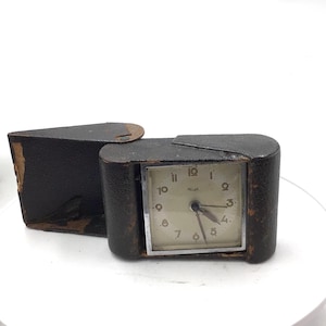 May include: A vintage travel alarm clock with a square white face, black numerals, and thin hands, housed in a dark brown textured leather case that opens on a hinge. The clock features a silver-toned metal frame around the dial.
