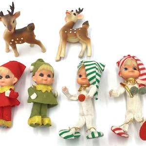 May include: A collection of six vintage-style figurines including two brown deer with white spots and four small dolls dressed in festive outfits of red, green, and white, featuring pointed hats, textured fabric clothing, and gold trim accents.