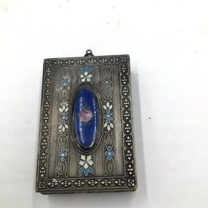 May include: A rectangular silver-toned metal case featuring an intricate engraved border and a central oval inlay. The inlay is deep blue enamel with a small pink rose and green leaves. Additional white and blue floral enamel accents decorate the surface.