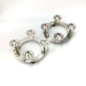 May include: Two silver-colored candle holders. Each circular holder has five candle cups. The holders are made of a shiny metal and sit on a white surface. The design is simple and modern, suitable for various decor styles.