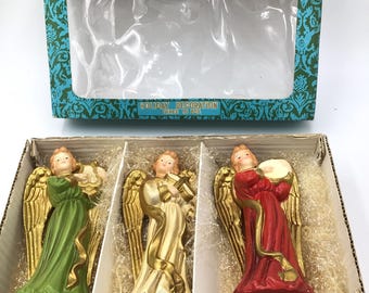 Puede incluir: Un conjunto de tres figuras de ángeles vintage en una caja. Los ángeles están hechos de yeso y están pintados en dorado, verde y rojo. Cada ángel sostiene un instrumento musical. La caja es azul y verde con las palabras "Happy Time" y "Decoración navideña" impresas en ella.