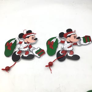 May include: Two flat wooden ornaments shaped like Mickey Mouse dressed in a red and white Santa suit. Each figure carries a green sack with a red bow and a small green gift box. A red string with a wooden bead hangs from the bottom of each ornament.
