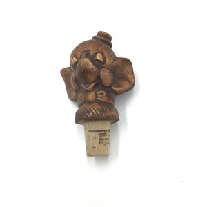May include: A brown ceramic bottle stopper shaped like a clown with a hat and a cork base.