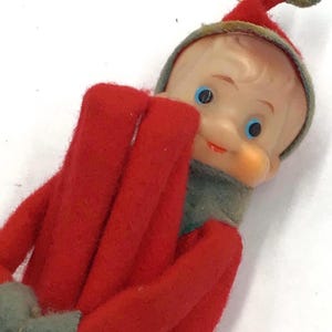 May include: A vintage-style elf figurine featuring a moulded plastic face with blue eyes and a red painted mouth. The elf wears a red felt suit with a matching pointed hat, grey felt collar, and grey felt mittens, with a gold hanging string.