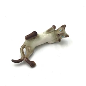 Op de afbeelding: Een keramiek beeldje van een Siamese kat die op zijn zij ligt met zijn poten omhoog. De kat is wit met bruine markeringen en heeft blauwe ogen.