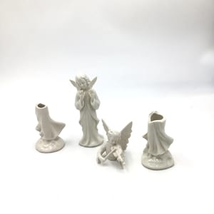 May include: Four white ceramic angel figurines. One angel is playing a violin, two are standing, and two are small candle holders. The angels have wings and are wearing robes. The figurines are on a white background.