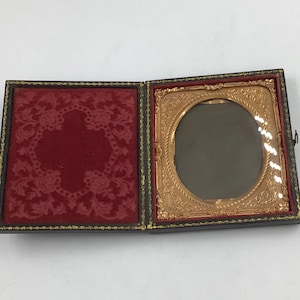 May include: An antique hinged case featuring a dark exterior with gold trim. The left side contains a deep red velvet lining with an embossed floral pattern. The right side holds an ornate gold-toned metal frame with an oval opening.