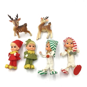 May include: A collection of vintage Christmas ornaments, including two brown reindeer figurines with white spots and four elf dolls in red, green, and white outfits. The elf dolls have blonde hair and are wearing hats and shoes.