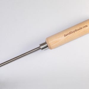 Può includere: Un utensile per la lavorazione del legno con un manico in legno chiaro e un'asta metallica. Il manico riporta la scritta "davidlloydtools.com". L'utensile ha una lama affilata e angolata all'estremità.