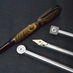 Puede incluir: Tres herramientas de metal con puntas de diferentes formas, incluyendo una cuadrada, una redonda y una puntiaguda. Las herramientas se utilizan para tornear madera. También se ve en la imagen una pluma de madera con un patrón de veta de madera negro y marrón.