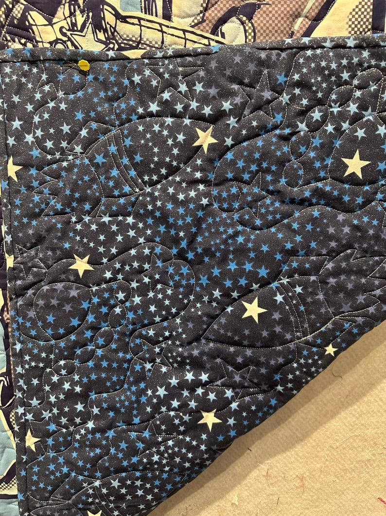 May include: A dark blue quilted fabric featuring a repeating pattern of light blue and cream-colored stars. The textile displays intricate stitching that outlines various shapes, including rocket ships and celestial motifs, across the surface.
