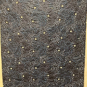 May include: A rectangular quilted fabric blanket featuring a dark navy blue background with a repeating pattern of light blue and yellow stars, along with subtle outlines of rocket ships and planets stitched into the fabric texture.