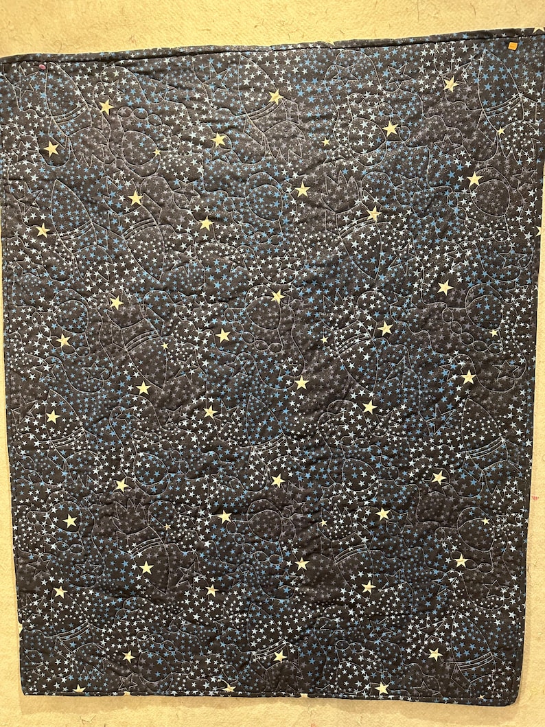 May include: A rectangular quilted fabric blanket featuring a dark navy blue background with a repeating pattern of light blue and yellow stars, along with subtle outlines of rocket ships and planets stitched into the fabric texture.