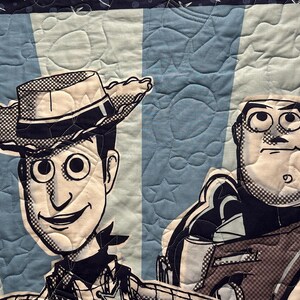 May include: A quilted fabric panel featuring a black and white graphic print of Woody and Buzz Lightyear. The background consists of alternating light and medium blue vertical stripes with a quilted star and circle pattern throughout the textile.