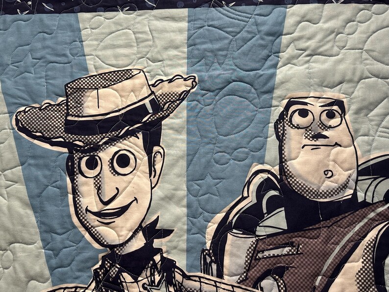 May include: A quilted fabric panel featuring a black and white graphic print of Woody and Buzz Lightyear. The background consists of alternating light and medium blue vertical stripes with a quilted star and circle pattern throughout the textile.