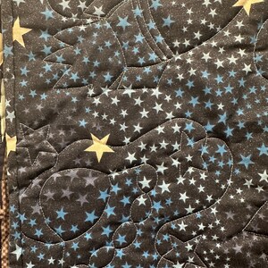 May include: A quilted fabric featuring a dark background with a dense pattern of small light blue and white stars. The quilt is stitched with curved lines that outline shapes of rockets and planets, accented by larger, solid gold-colored stars.