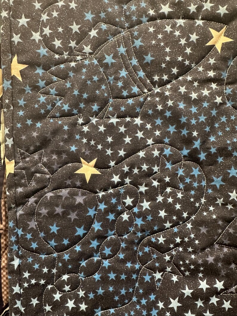 May include: A quilted fabric featuring a dark background with a dense pattern of small light blue and white stars. The quilt is stitched with curved lines that outline shapes of rockets and planets, accented by larger, solid gold-colored stars.