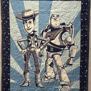 May include: A rectangular quilted blanket featuring a graphic print of two cartoon characters, a cowboy and a space ranger, set against a light blue sunburst background. The quilt has a dark blue border decorated with white stars and planet motifs.