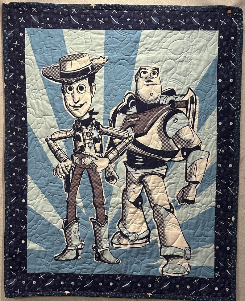 May include: A rectangular quilted blanket featuring a graphic print of two cartoon characters, a cowboy and a space ranger, set against a light blue sunburst background. The quilt has a dark blue border decorated with white stars and planet motifs.